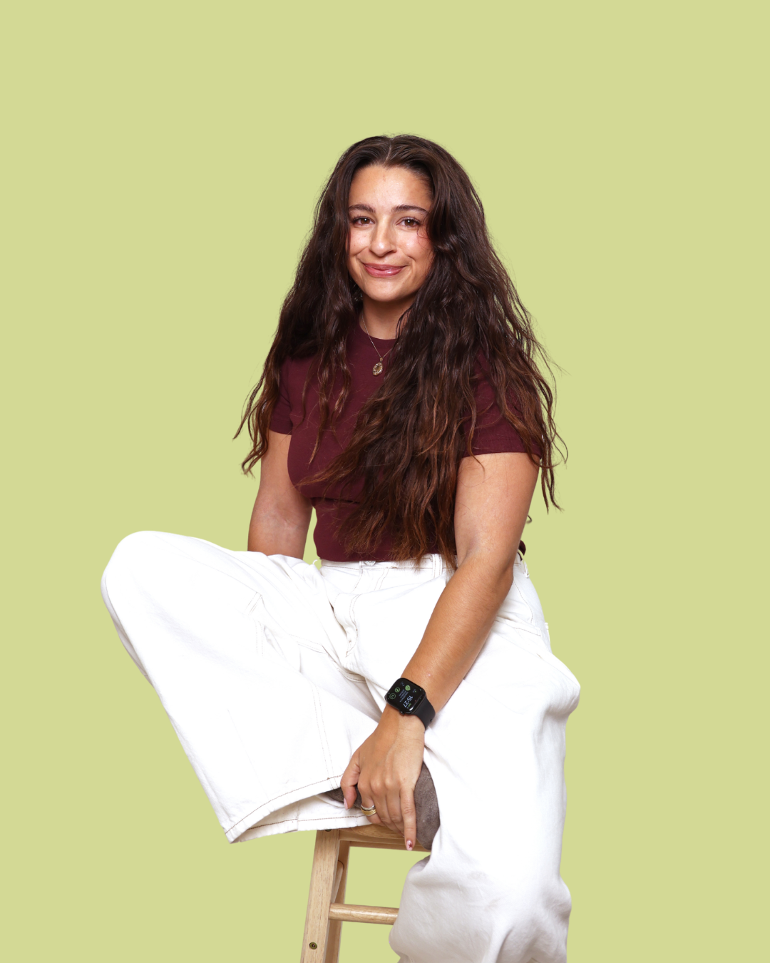 A woman with long dark hair sits on a wooden stool against a solid pale yellow-green background. She is wearing a dark burgundy short-sleeve top and white wide-leg pants.