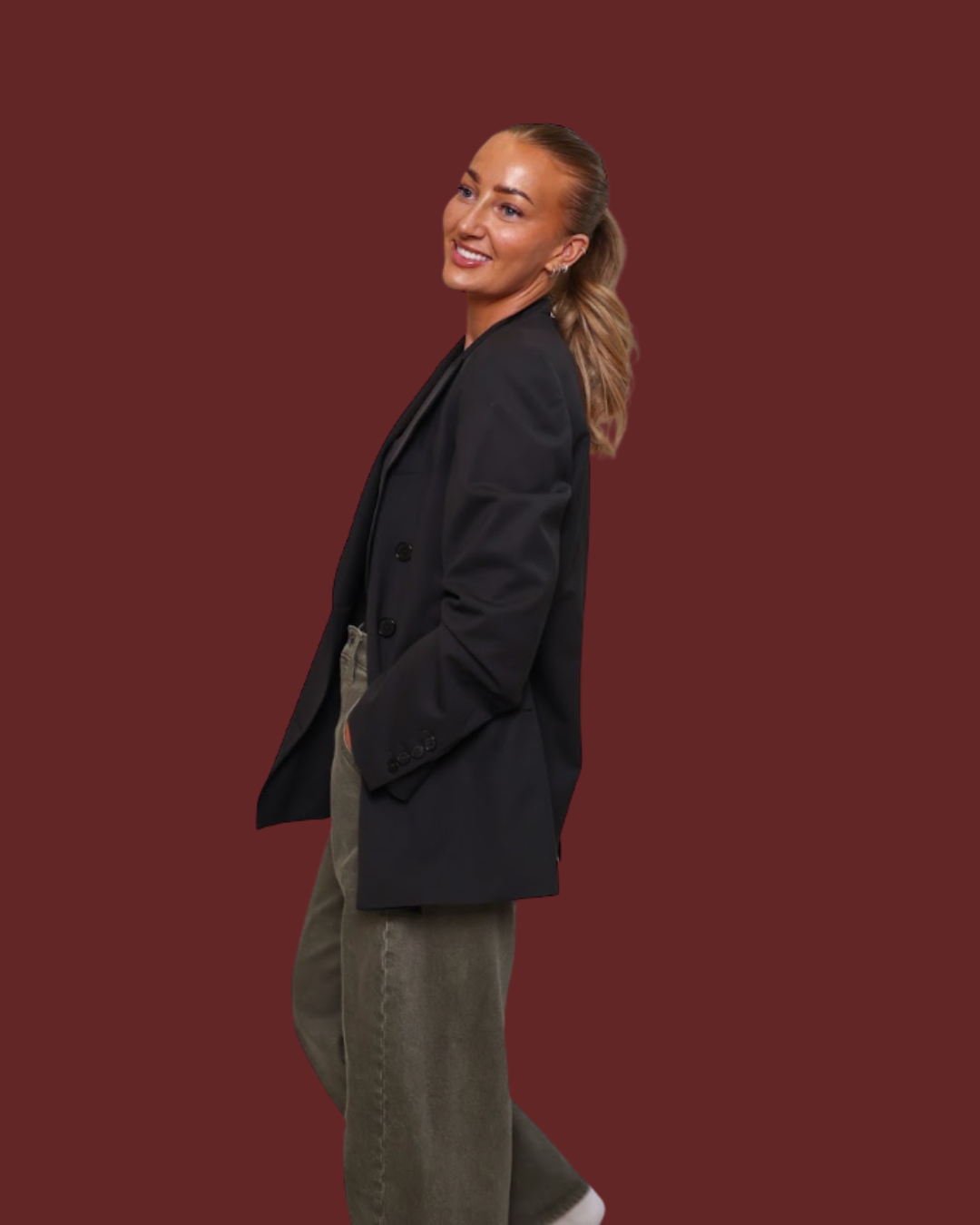 A woman posing in a black double-breasted blazer and olive green wide-leg trousers against a solid dark red background.